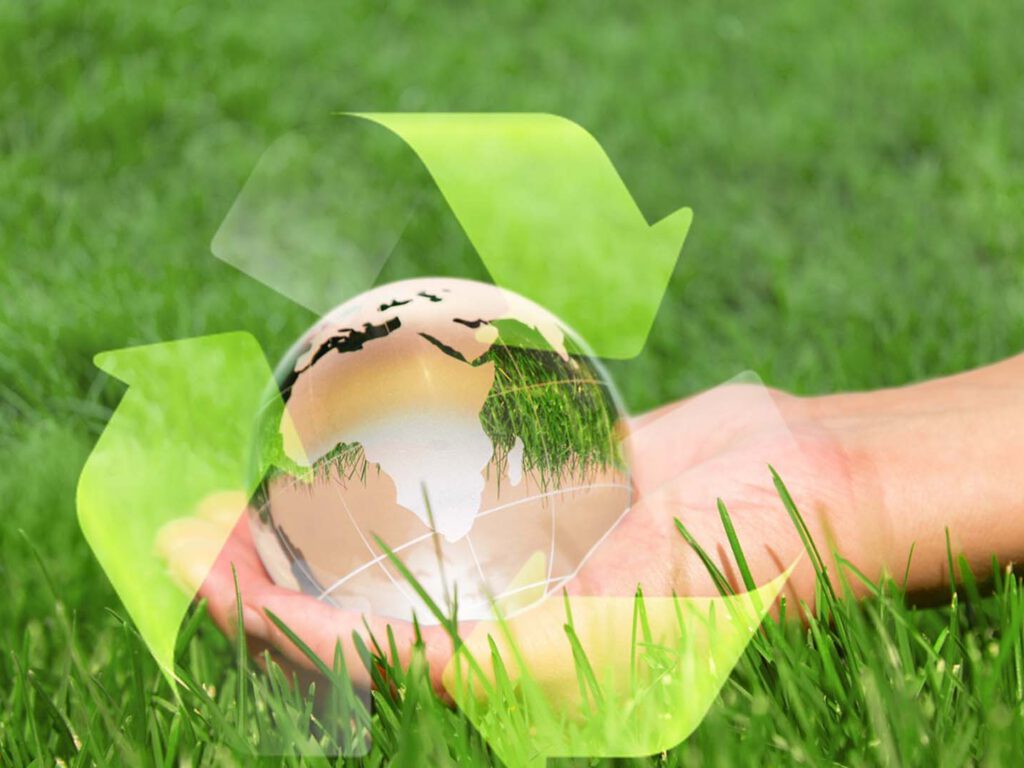 Eine Hand liegt mit dem Handrücken auf dem Rasen und hält eine Weltglaskugel. Um die Kugel sind die Pfeile der Kreislaufwirtschaft dargestellt